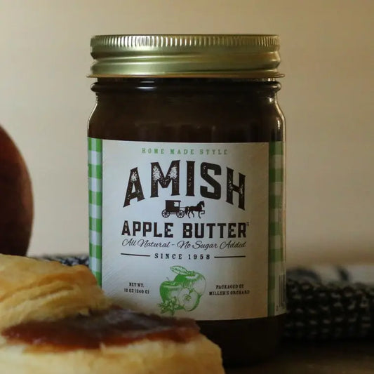 A glass jar of Amish Apple Butter with a gold metal lid and a white label with green gingham accents, shown with a pastry in the foreground on a neutral beige background.
