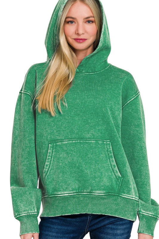 A woman wearing a forest green acid-wash pullover hoodie with a front kangaroo pocket, paired with blue jeans, against a plain white background.