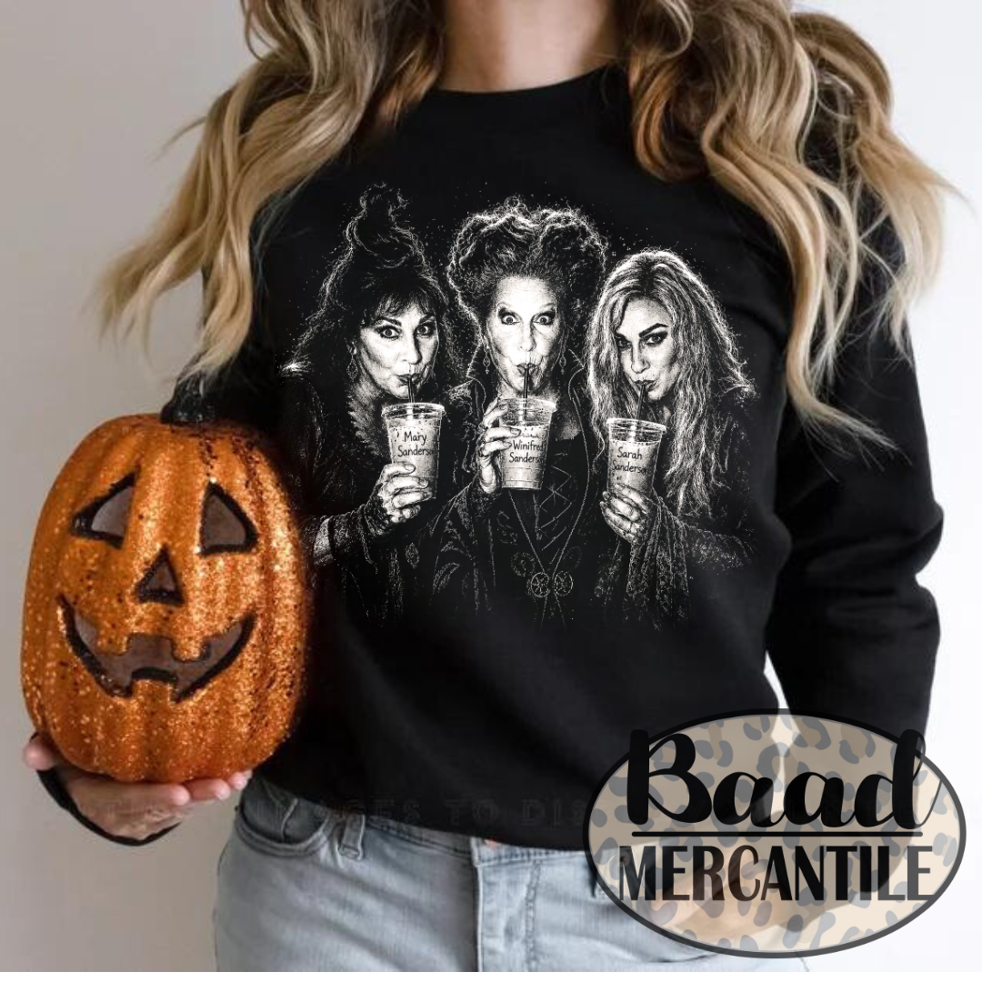 A woman wearing a black long-sleeve graphic sweatshirt with a white illustration of three witches holding cups, holding a glittery orange jack-o'-lantern. A 'Baad Mercantile' watermark is visible in the bottom right corner.